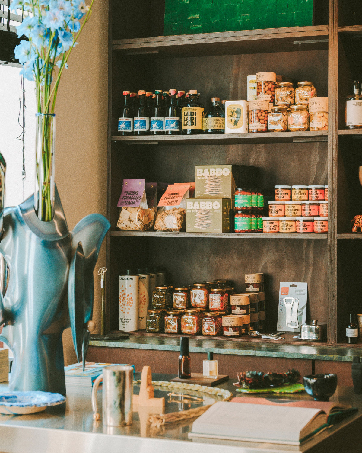 A styled interior scene showing shelves stocked with jars, bottles, and packaged food products, alongside a large blue vase with flowers on a counter in the foreground.
