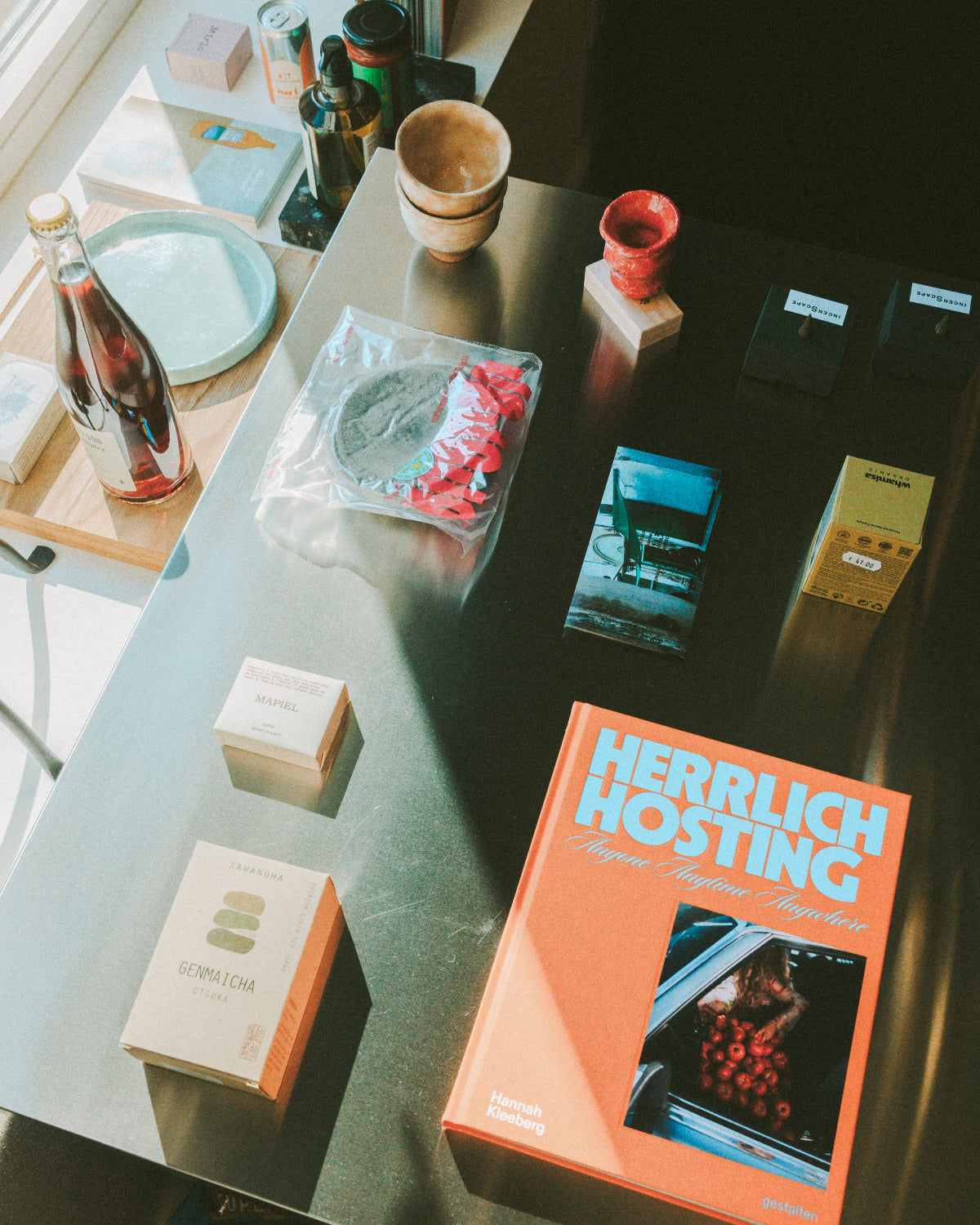 A styled overhead shot of a dark tabletop with several products arranged on it, including a large orange book titled 'HERRLICH HOSTING', small product boxes, a bottle, bowls, and other objects in a sunlit interior setting.